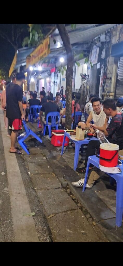 Locals sit on small plastic stools on a sidewalk to enjoy a meal